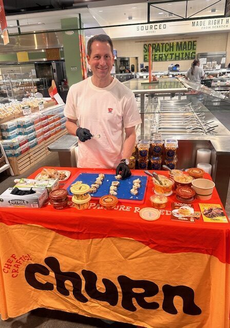 A man in a white T-shirt and black gloves stands behind a table with food samples and dips in a grocery store, with a "churn" banner in front.