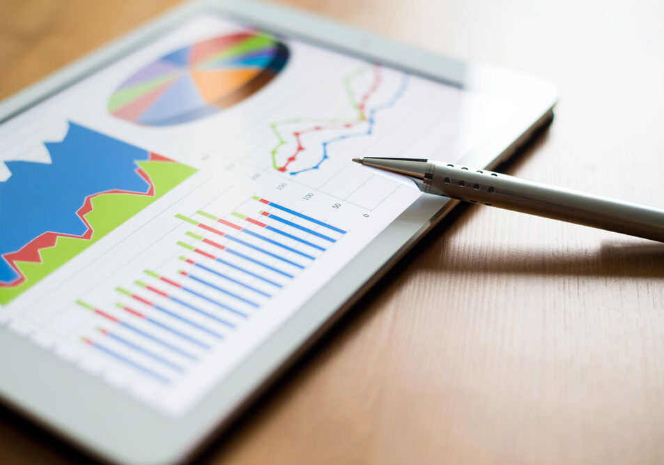 Tablet displaying various financial graphs and charts, including bar graphs, line charts, and a pie chart, with a pen resting nearby on a wooden surface.