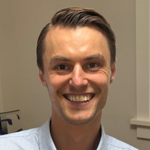 Headshot of Jonathan Coppel, a man with short brown hair and a light blue collared shirt smiles at the camera in an indoor setting with beige walls.