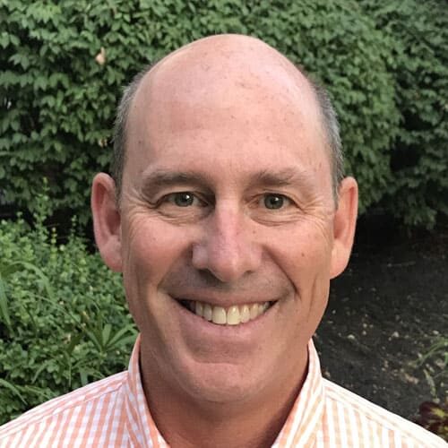 Headshot of Rich Tashman, a middle-aged man with a bald head and striped shirt smiles at the camera, standing outdoors in front of leafy green bushes.