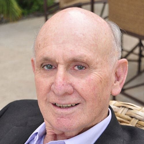 Headshot of Stan Tashman, an older man with a bald head and light skin, wearing a black jacket and light-colored shirt, sits outdoors and looks at the camera.