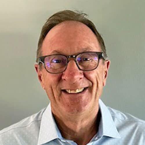 Headshot of Ty Olson, a middle-aged man wearing glasses and a light blue collared shirt smiles at the camera in front of a plain light-colored background.