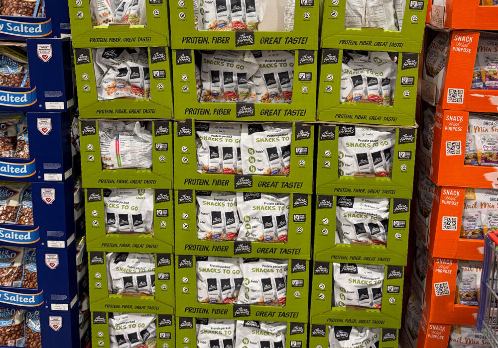 Boxes of "Snacks To Go" protein and fiber snack packs stacked on a pallet in a warehouse store, next to displays of other snack products.