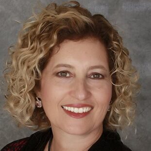 Headshot of Vicki Tashman, a woman with short curly blonde hair smiling at the camera, wearing earrings and a dark patterned top, against a gray background.